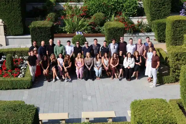 Group portrait of coworkers posing in a formal garden courtyard with trimmed hedges and flowers.
